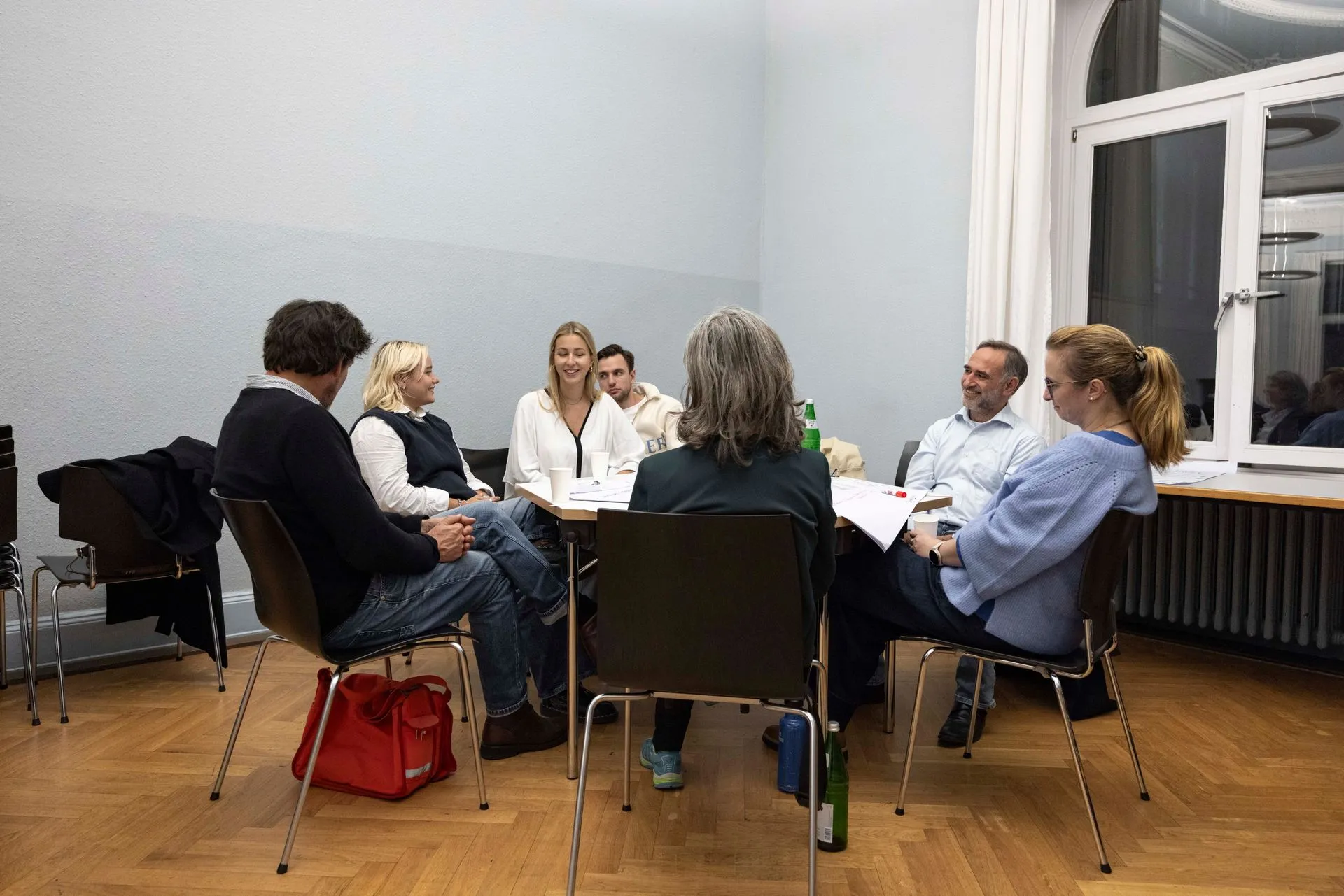 Eine Gesprächsrunde: neun Menschen unterschiedlichen Alters sitzen im Kreis um einen Tisch, einige beugen sich vor.